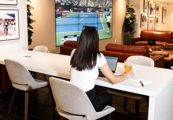 Young woman sitting in the WorkHUB at the Adelaide Club with her laptop and a beer, working while watching sports on the big screen